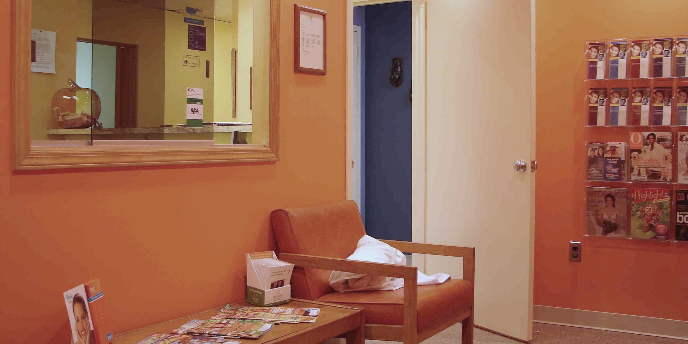 The waiting room at Sunbeam Dental: orange walls, an orange armchair and a table of magazines, a window through to the front desk and an open door to a blue hallway